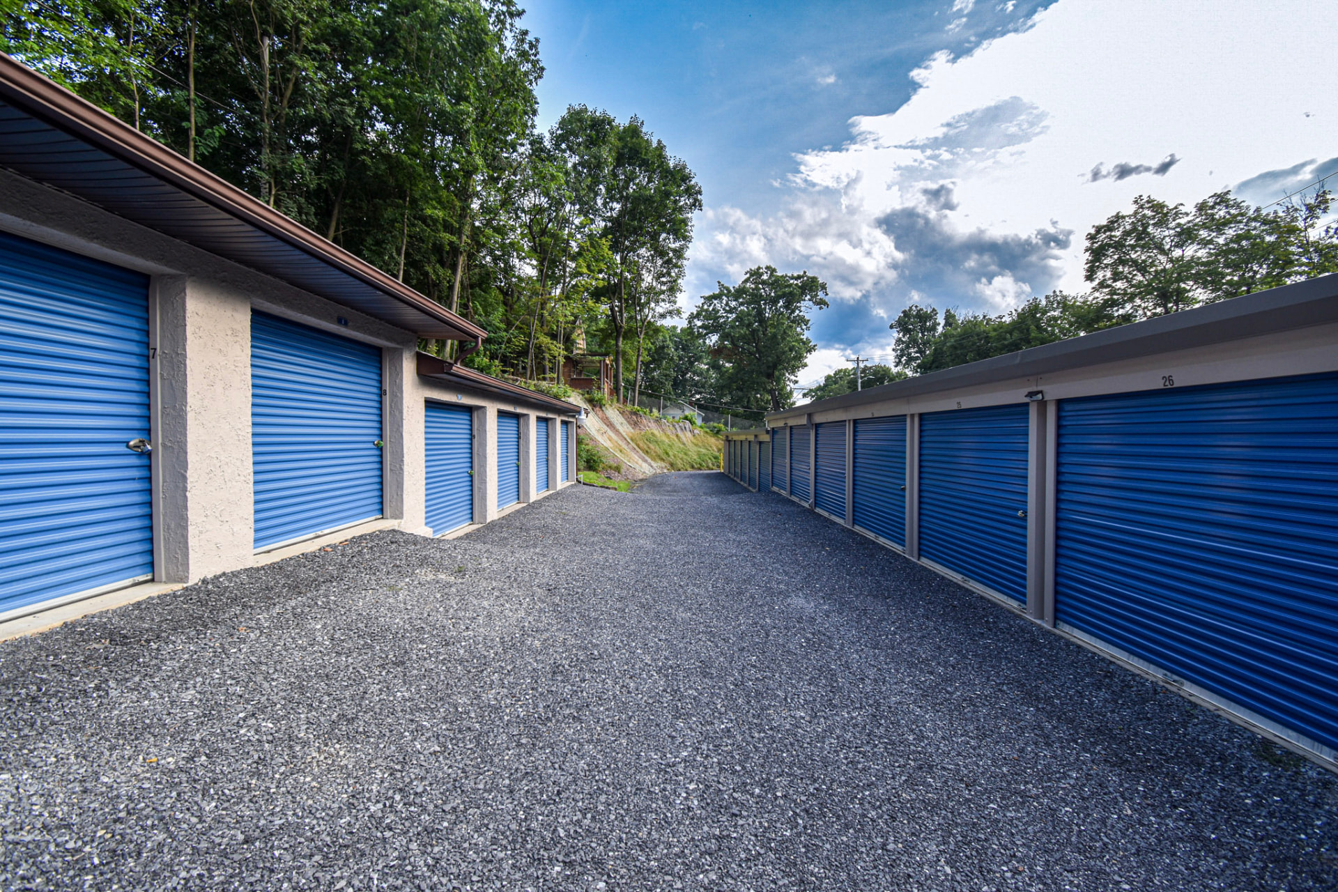 After: the same storage drive with new blue roll-up doors, clean walls, and fresh gravel.